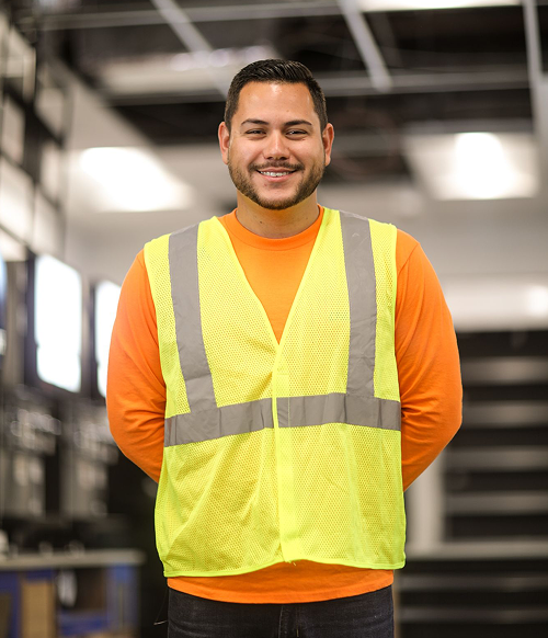 Smiling man wearing an orange shirt and a yellow reflective safety vest in an industrial setting.