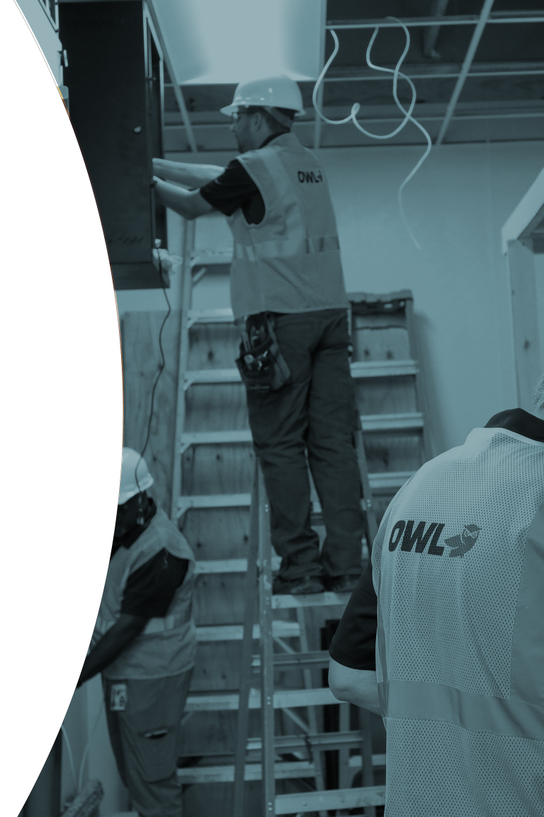 Three construction workers wearing hard hats and OWL vests working inside a building with ladders and exposed wiring.