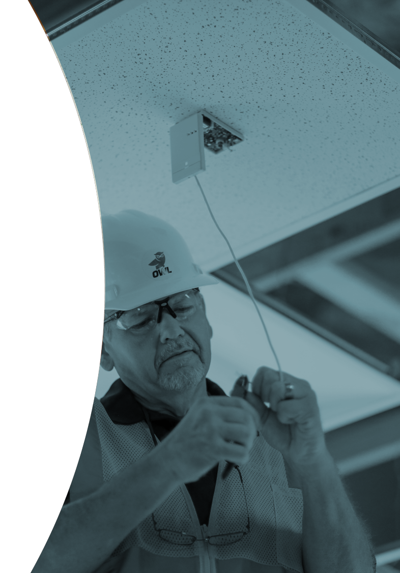 Construction worker wearing safety glasses and a hard hat repairing a glass break sensor on a ceiling panel.