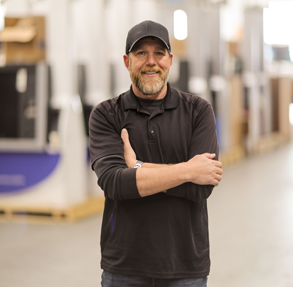 Smiling man with a beard and black cap wearing a black long-sleeve polo shirt standing with arms crossed in a warehouse.