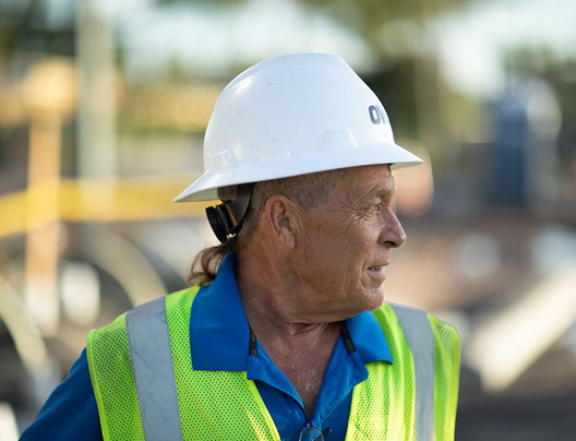 Two men, one in a construction vest and hard hat, and another in a blue shirt, discussing inside a building under construction with a red ladder in the background.