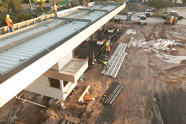 Construction site with workers assembling a large elevated steel structure over a dirt ground.