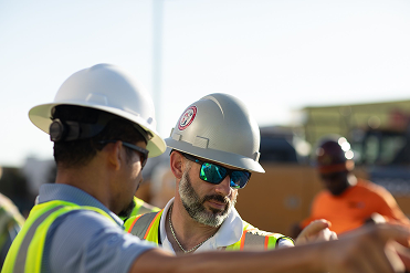 Two construction workers wearing helmets and safety vests, one with sunglasses, at a worksite.