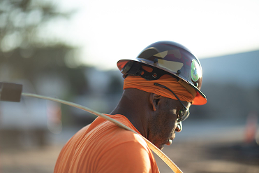 Construction worker wearing a camouflage hard hat, orange shirt, and safety glasses, looking down at a worksite.