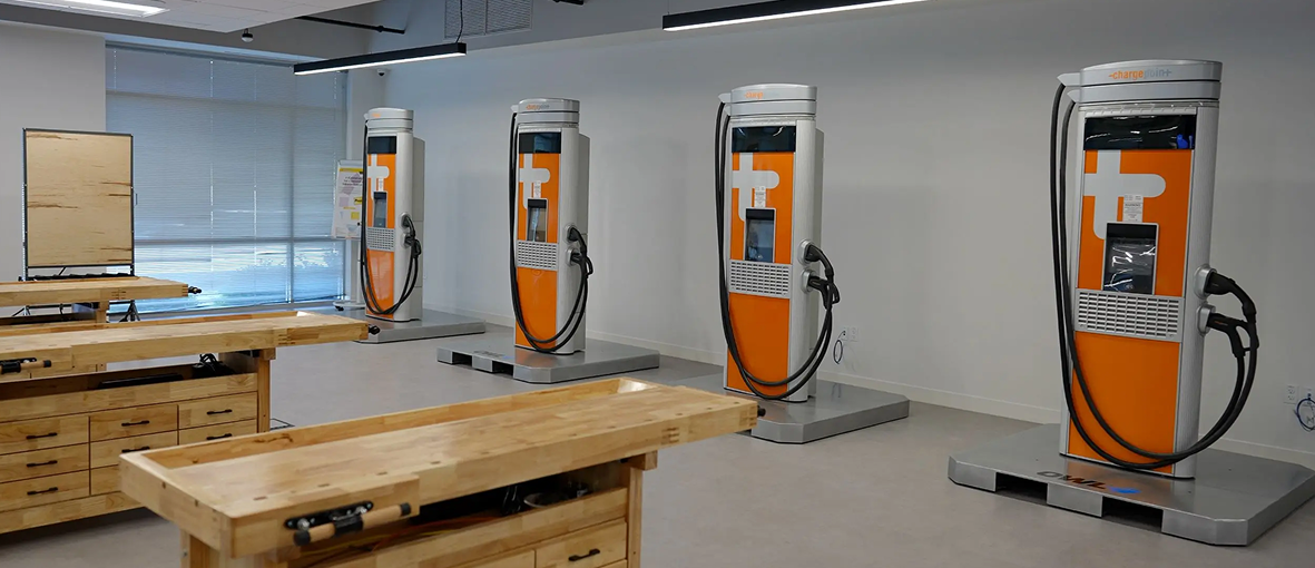 Indoor setup with four orange and silver electric vehicle charging stations aligned against a white wall, with wooden workbenches in the foreground.