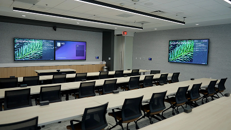 Modern classroom with multiple rows of black chairs and desks facing two large wall-mounted screens displaying digital content.