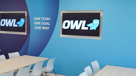 Modern meeting room with blue walls, a TV displaying the OWL logo with an owl icon, and the words 'ONE TEAM ONE GOAL ONE WAY' on the wall.