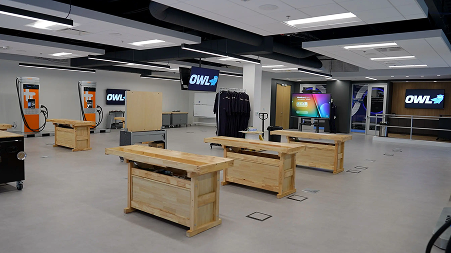 Spacious modern classroom with wooden tables, screens displaying 'OWL' logo, and orange charging stations on the left wall.