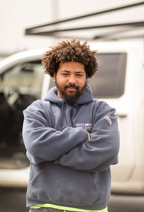 Man with curly hair and beard wearing a blue hoodie standing with arms crossed in front of a white vehicle.
