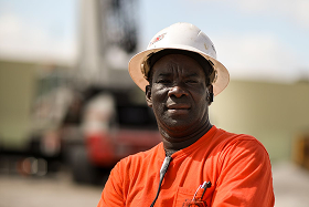 Construction worker wearing a white hard hat and orange shirt looking at the camera with a blurred industrial background.