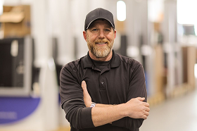 Smiling middle-aged man with a beard wearing a black cap and shirt, standing with arms crossed in a bright indoor workspace.