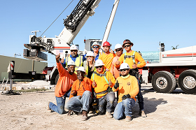 Group of diverse construction workers wearing helmets and safety gear posing cheerfully on a worksite with construction equipment in the background.