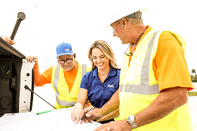 Two construction workers and a woman in a blue shirt reviewing documents on a construction site.
