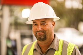 Man wearing a white construction helmet and yellow safety vest smiling outdoors.