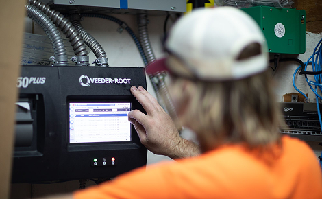 Person wearing a white cap and orange shirt interacting with a Veeder-Root device screen in a utility room.