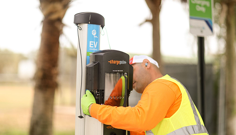 Worker in orange safety vest and gloves installing or inspecting an EV charging station outdoors near palm trees.