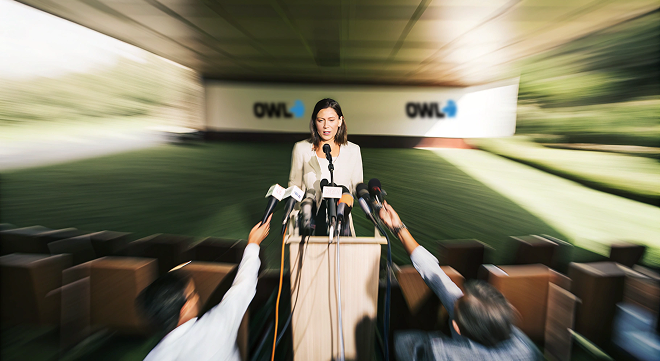 Woman speaking at a podium with multiple microphones, flanked by reporters holding out microphones.
