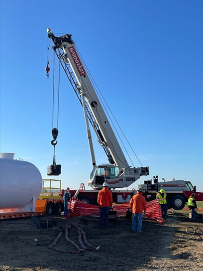 Construction site with a crane lifting a load, three workers in orange and yellow safety vests, and a large white tank on a trailer under a clear blue sky.