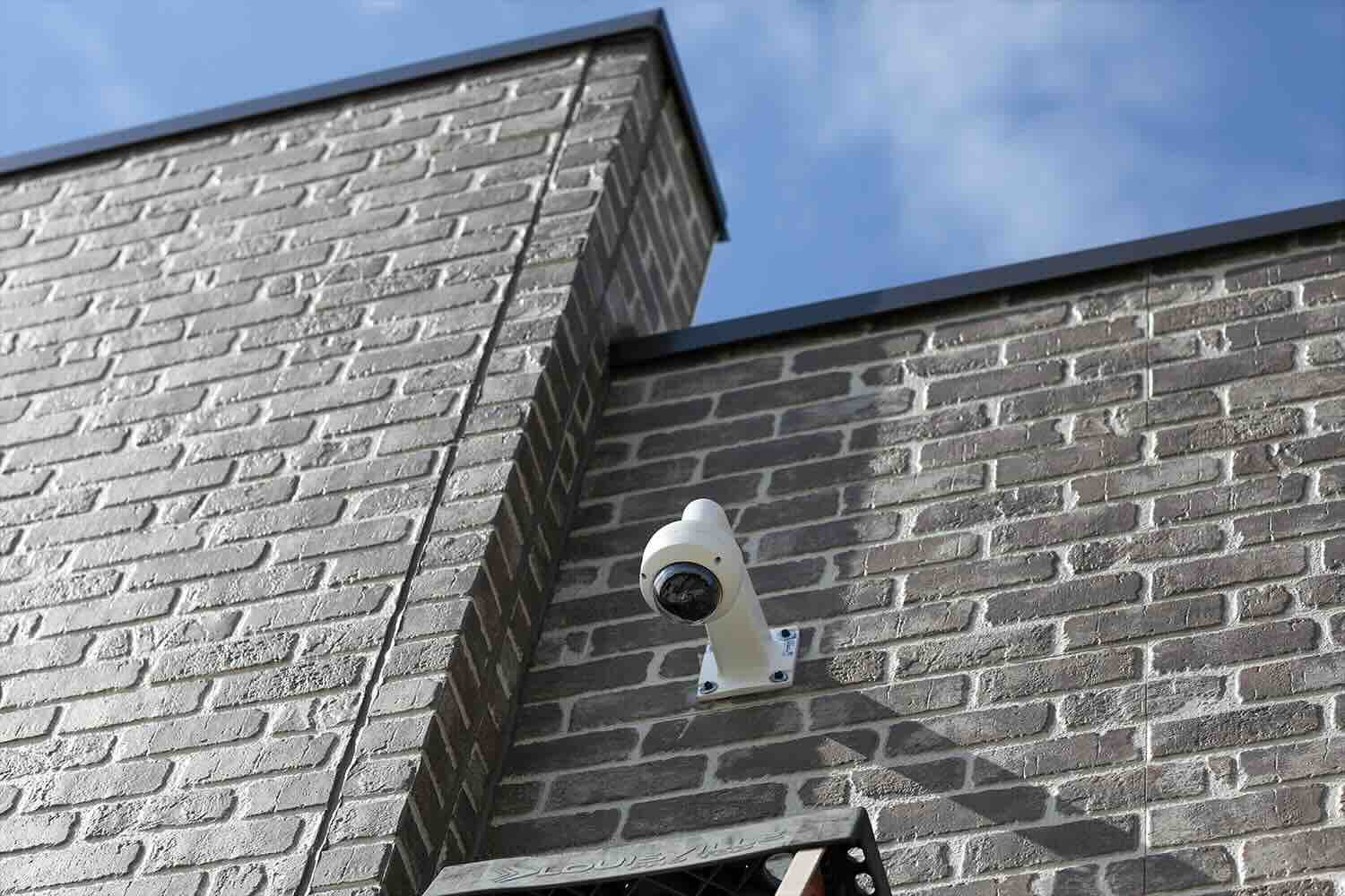 White surveillance camera mounted on a gray brick wall under a blue sky.