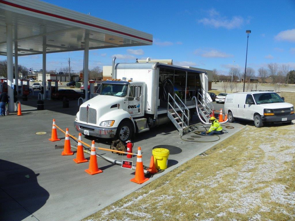 Worker in safety gear performing maintenance on underground fuel lines at a gas station, with a white service truck and safety cones around the work area.