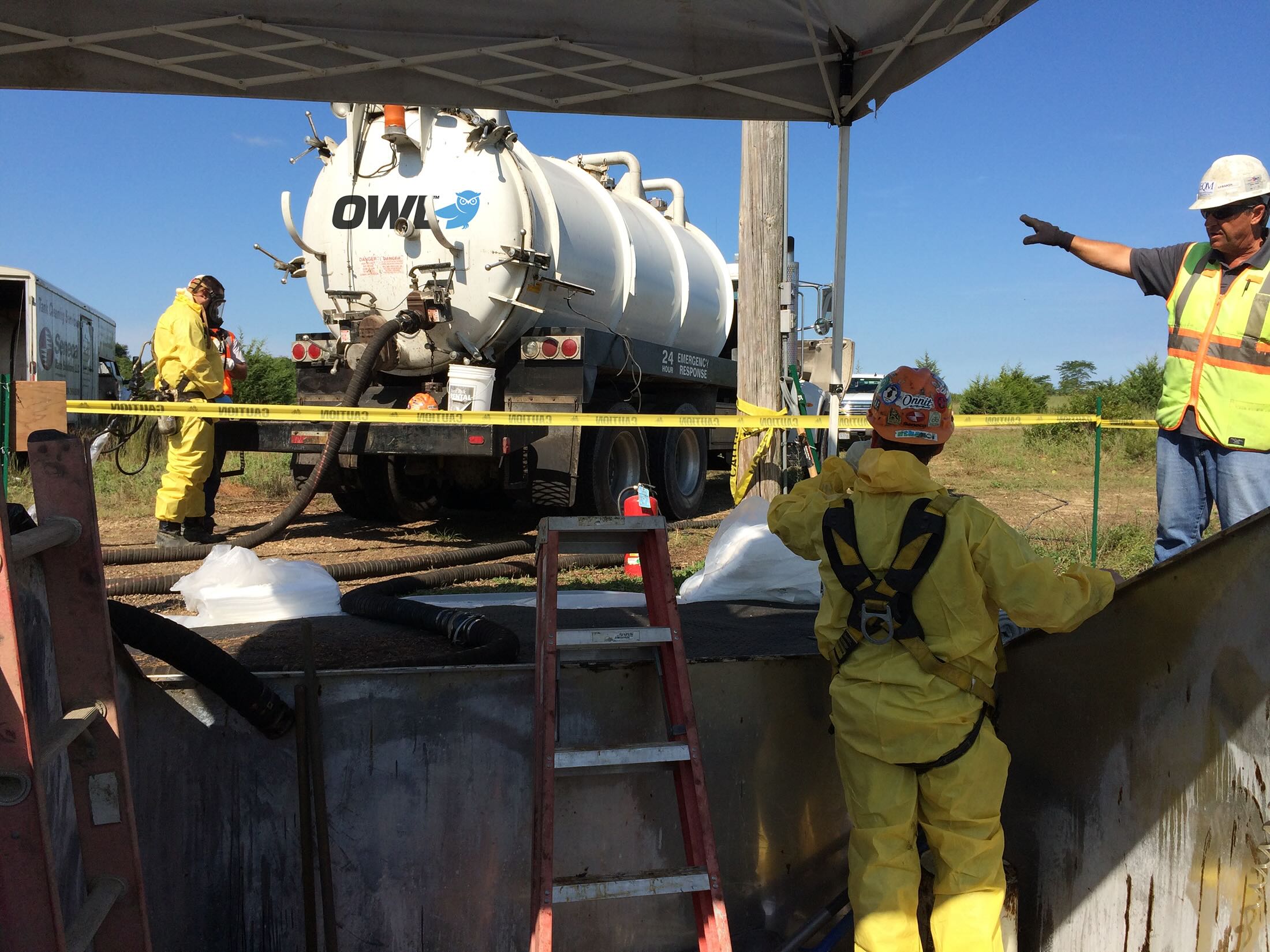 Workers in yellow protective suits at a confined space excavation site with an industrial vacuum truck and caution tape.
