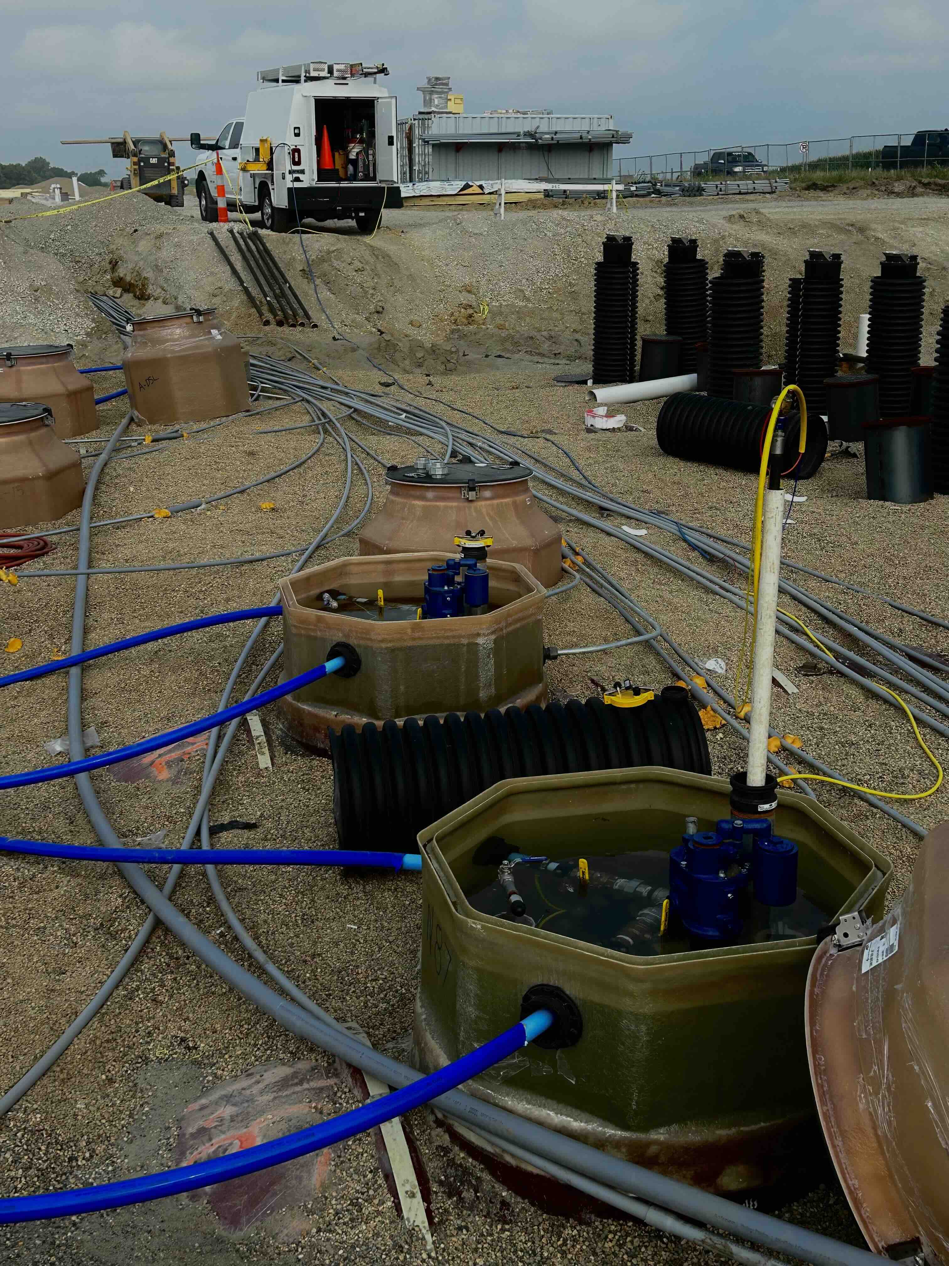 Construction site with large underground utility tanks connected by blue and gray pipes, a white utility truck, and stacked black corrugated pipes in the background.