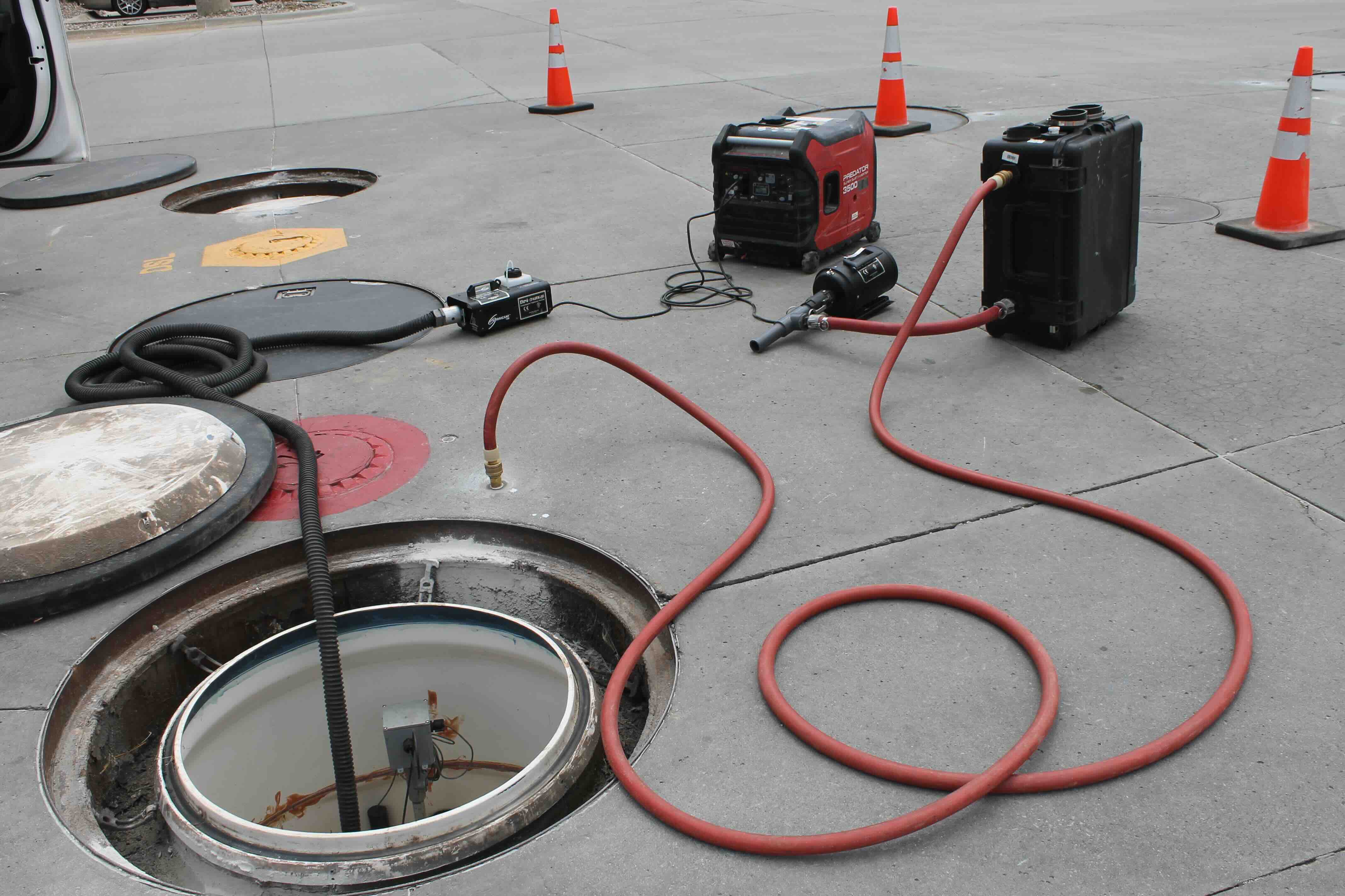 Open manholes on a concrete street with testing equipment, including a portable generator and connected hoses, surrounded by safety cones.