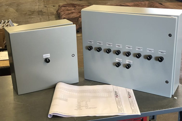 Two gray industrial control panels with multiple black knobs on a table alongside a folded technical schematic.