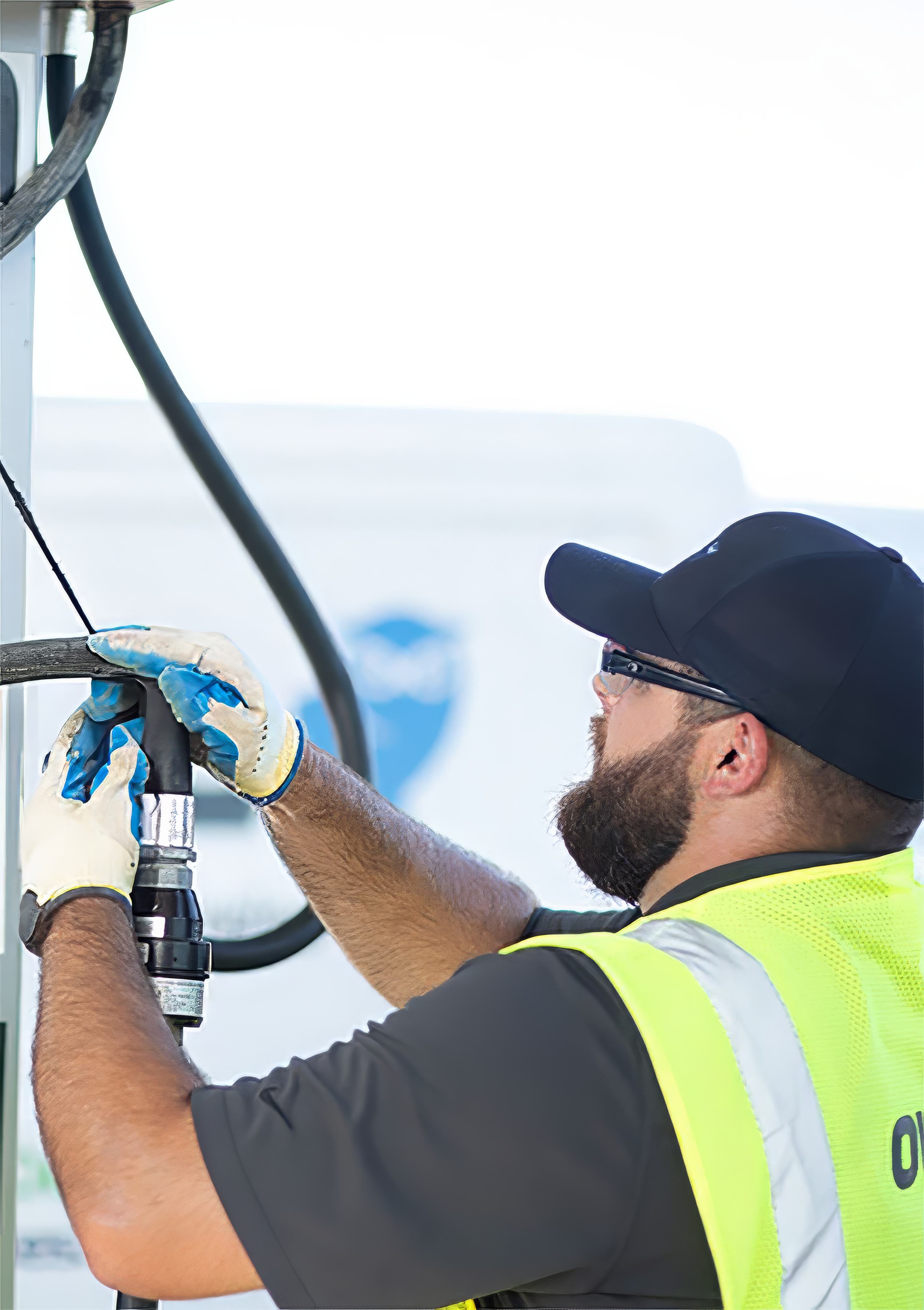 Worker wearing gloves, a cap, safety glasses, and a yellow vest handling a large hose or fuel nozzle.