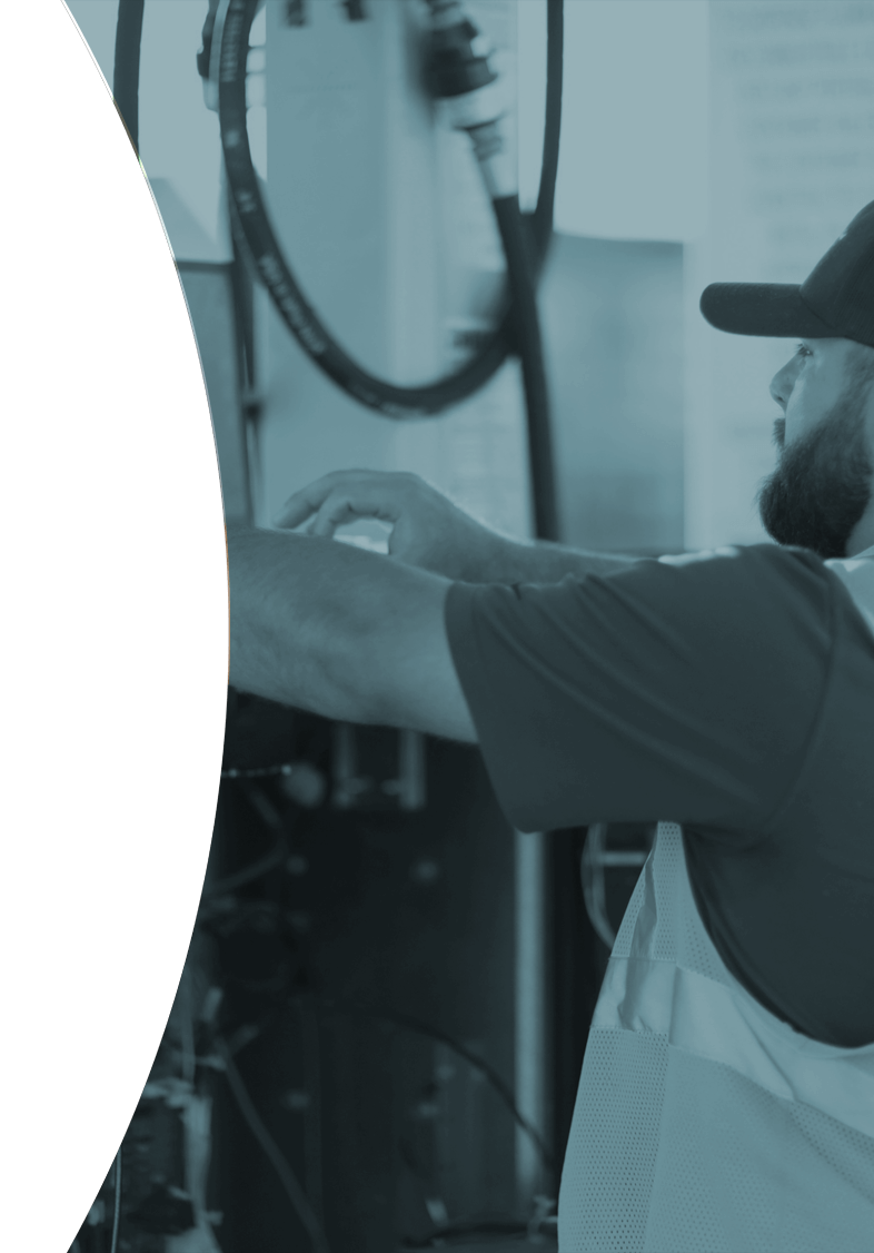 Worker in a cap and safety vest operating laptop at a fuel dispenser setting.