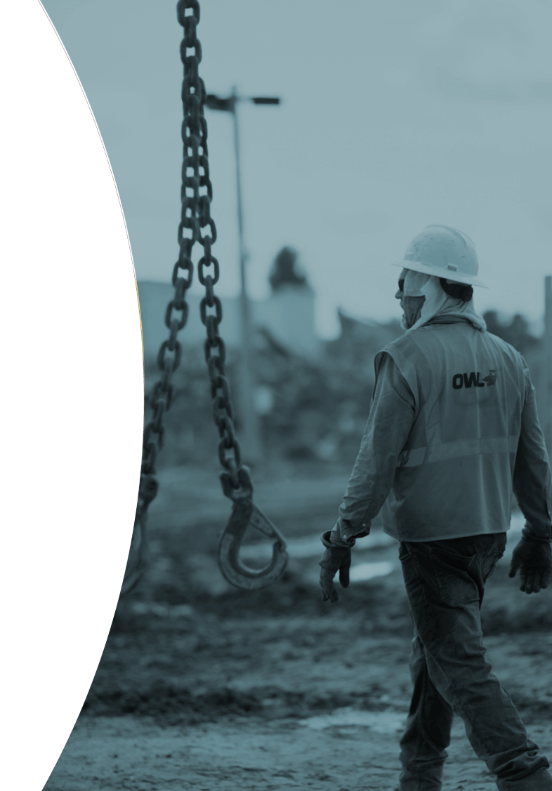 Construction worker wearing a hard hat and OWL vest walking near a hanging metal chain hook on a muddy site.