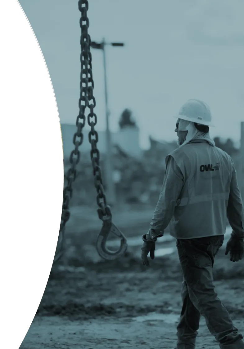 Construction worker wearing a hard hat and OWL vest walking near a hanging metal chain hook on a muddy site.