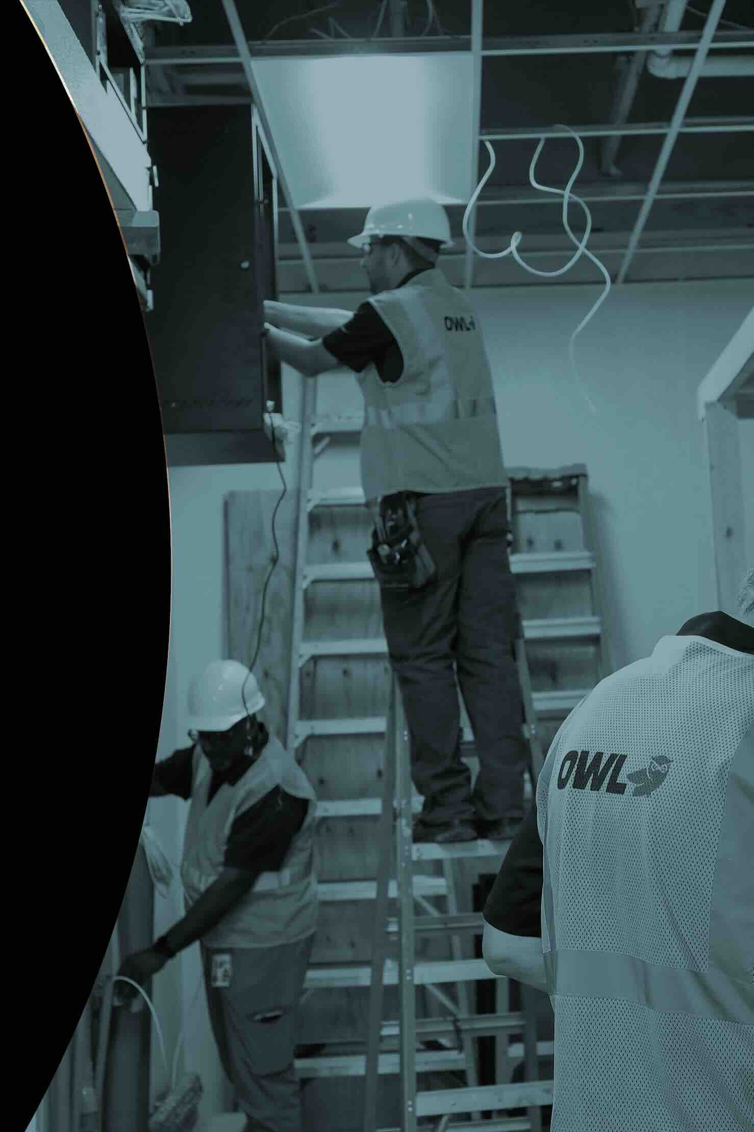 Three construction workers wearing hard hats and OWL vests working inside a building with ladders and exposed wiring.
