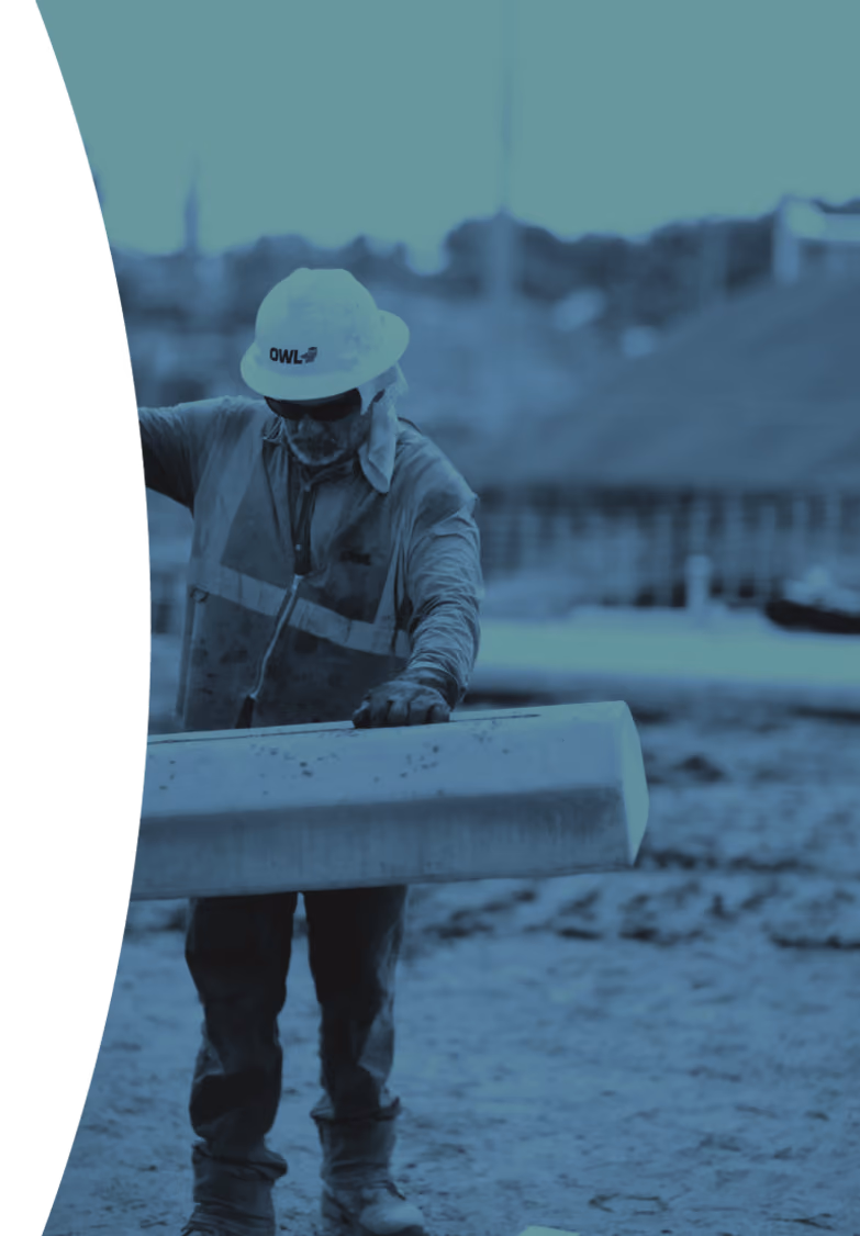 Construction worker wearing a hard hat and safety vest examining a concrete "deadman" beam on a job site.