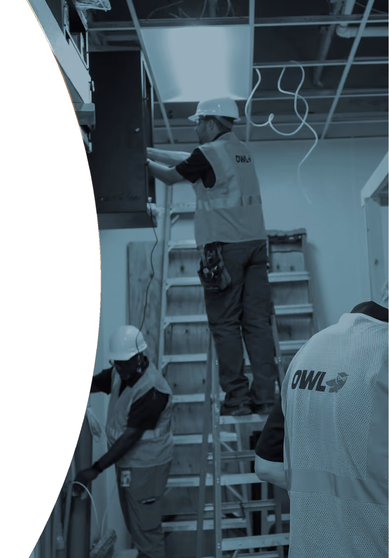 Three construction workers wearing hard hats and OWL vests working inside a building with ladders and exposed wiring.