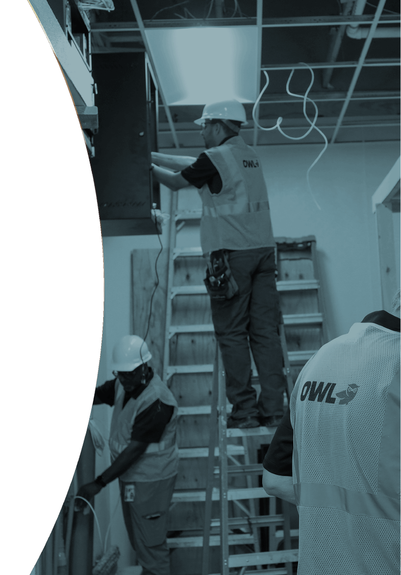 Three construction workers wearing hard hats and OWL vests working inside a building with ladders and exposed wiring.
