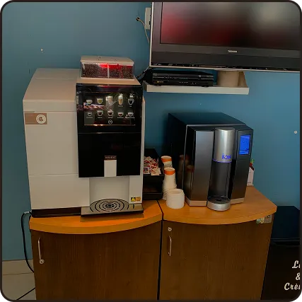 Coffee station with a large coffee machine, a smaller touchscreen coffee dispenser, and disposable cups on a wooden counter under a wall-mounted TV.