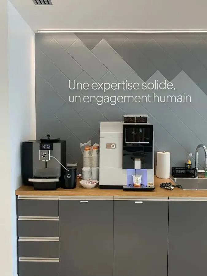 Coin café avec machines à café, gobelets, évier, et mur gris avec texte Une expertise solide, un engagement humain.