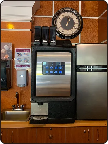 Machine à café automatique avec écran tactile, horloge murale et évier dans une cuisine.