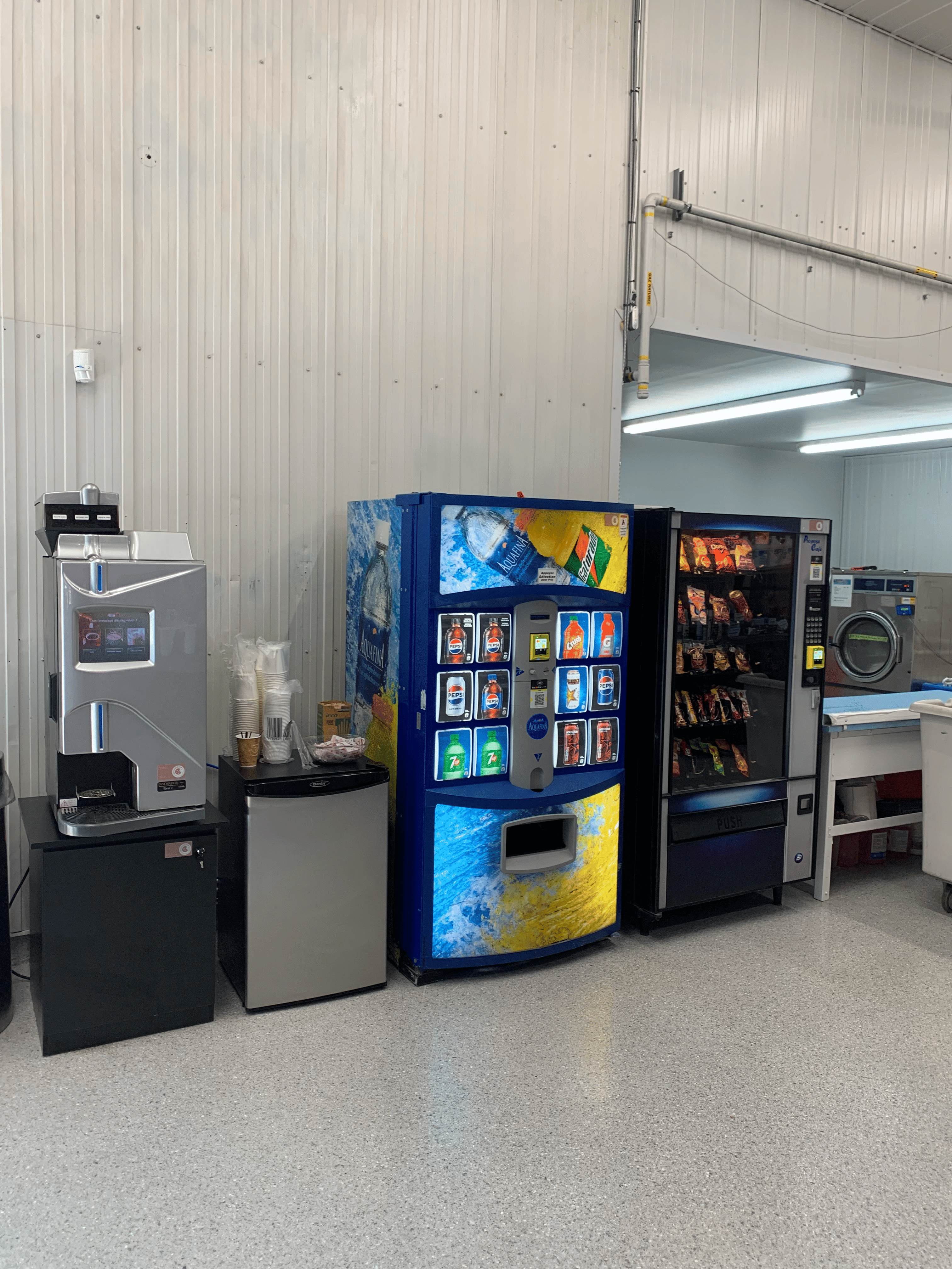Distributeurs automatiques de boissons et collations alignés dans une salle avec machine à café et mini-réfrigérateur.