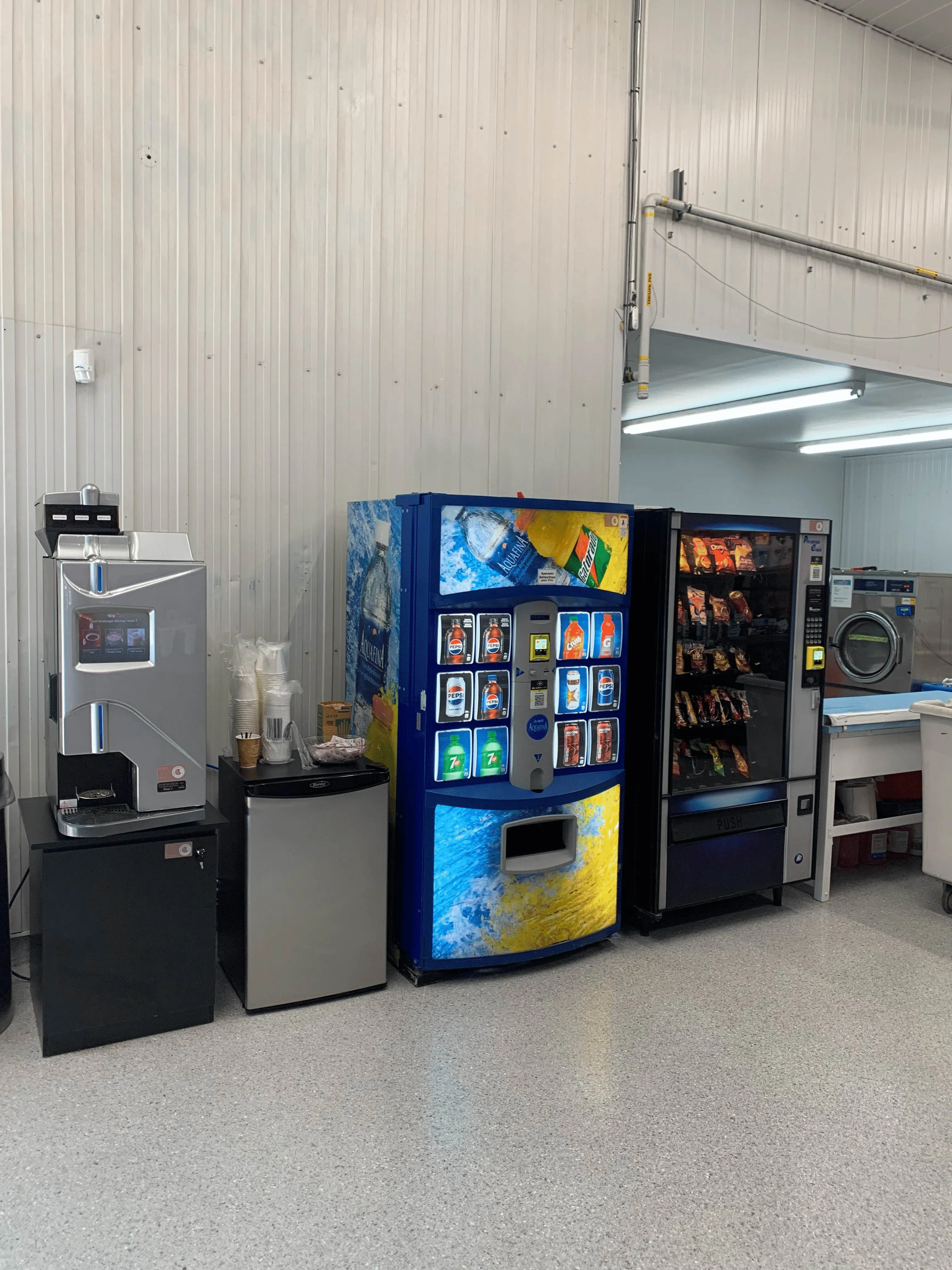 Distributeurs automatiques de boissons et collations alignés dans une salle avec machine à café et mini-réfrigérateur.