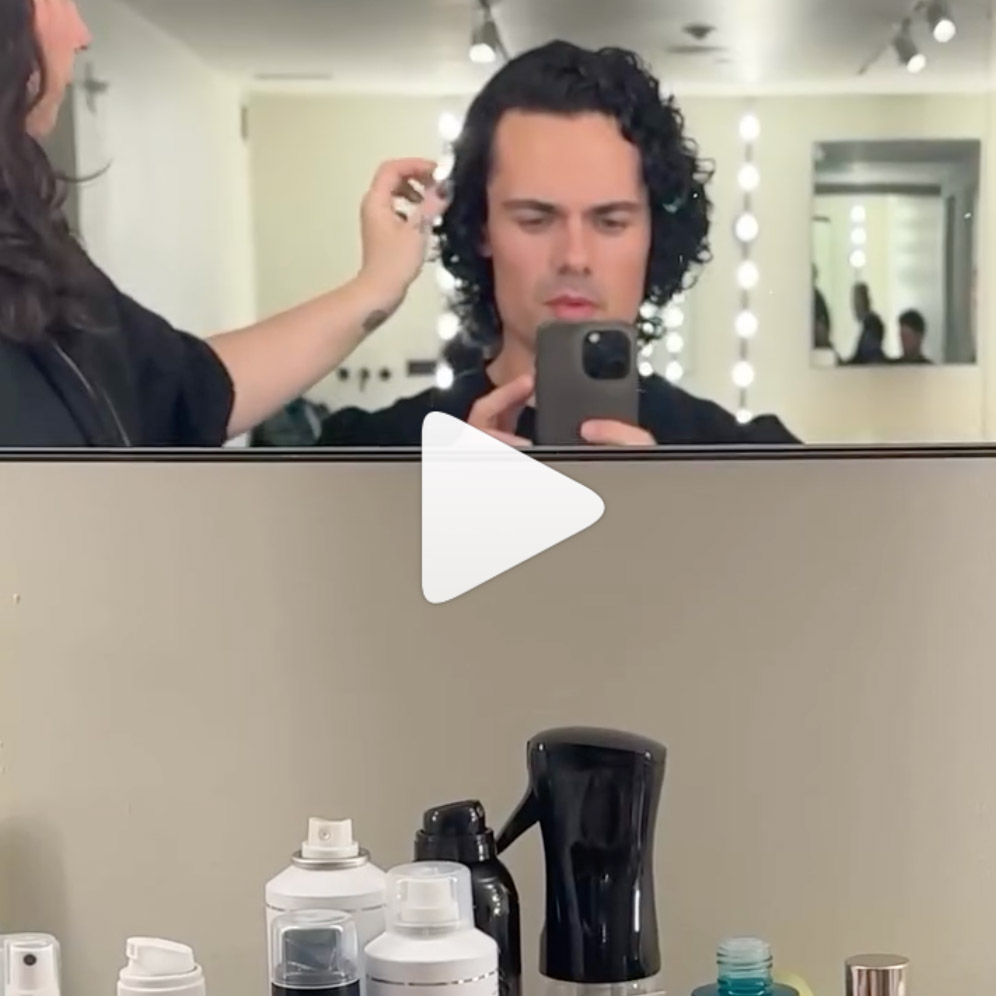 Man with curly dark hair taking a mirror selfie while a stylist adjusts his hair in a well-lit salon.