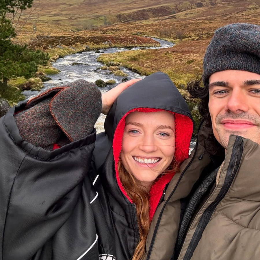 Smiling couple wearing warm jackets and hats taking a selfie outdoors near a flowing stream in a mountainous landscape.