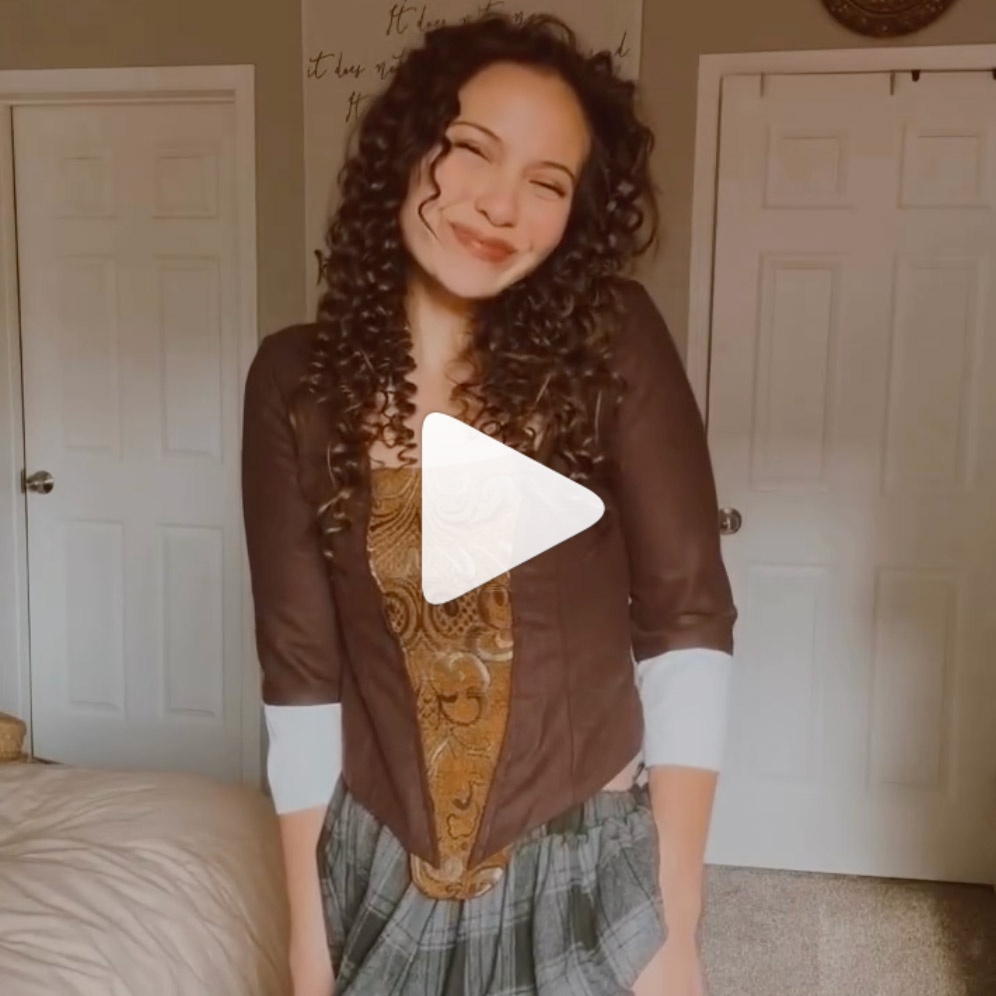 Smiling woman with curly hair wearing a brown and patterned top and plaid skirt standing indoors.