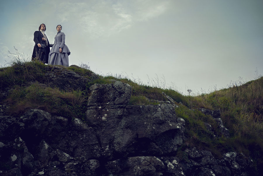 Man and woman in period clothing standing on a grassy rocky hill under a cloudy sky.