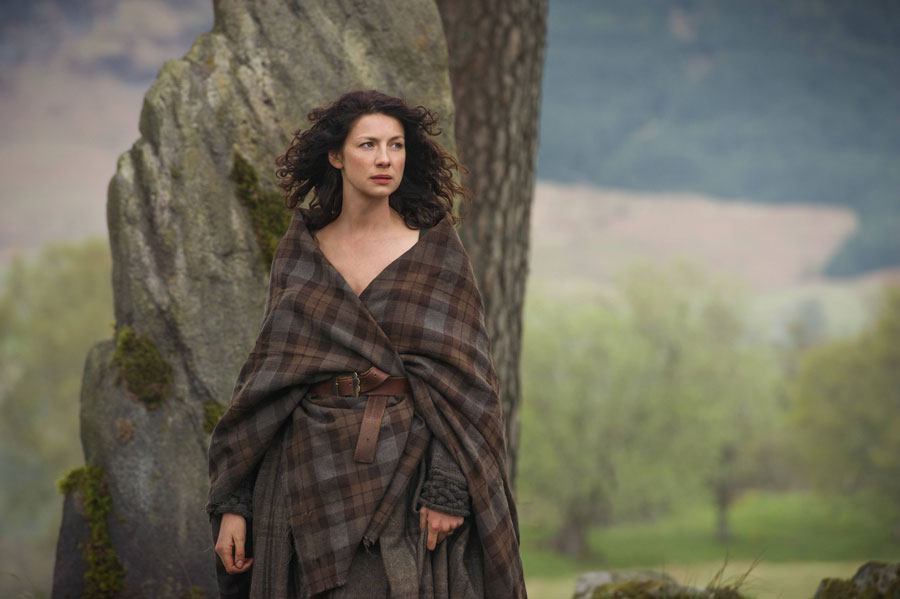 Woman with curly dark hair wrapped in a brown plaid shawl standing outdoors near a large rock with a forested landscape in the background.