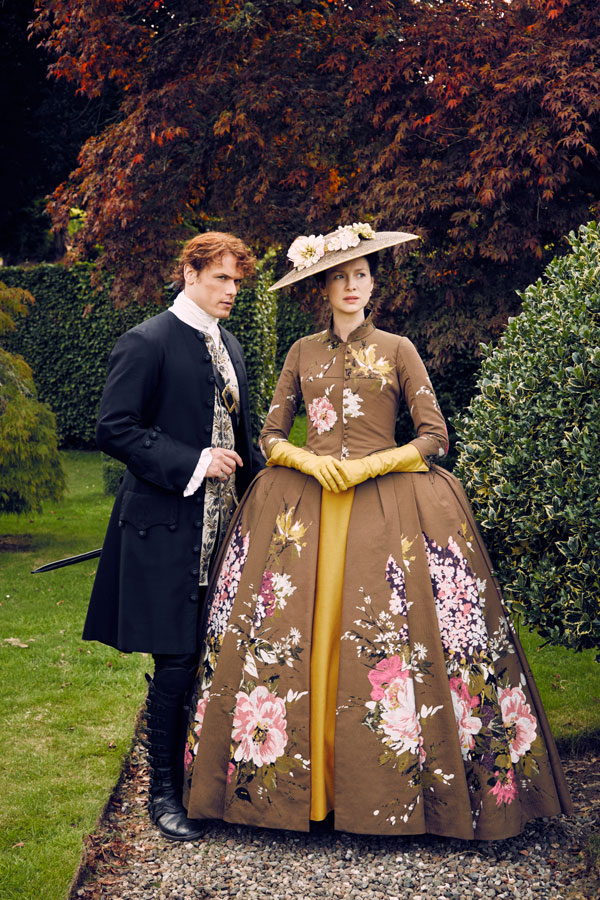 A man and woman in elaborate 18th-century style attire stand outdoors; the woman wears a wide floral dress with a matching hat and yellow gloves, while the man wears a dark coat with white cravat.