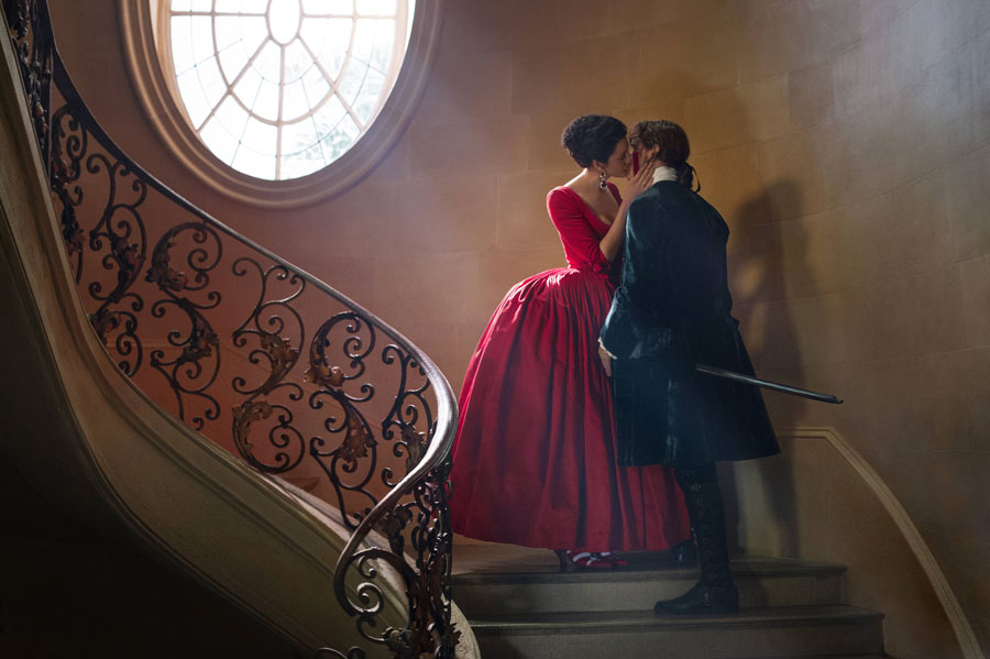 A woman in a red historical gown embraces and kisses a man in dark period clothing holding a sword on a curved staircase with an ornate railing and a large round window.