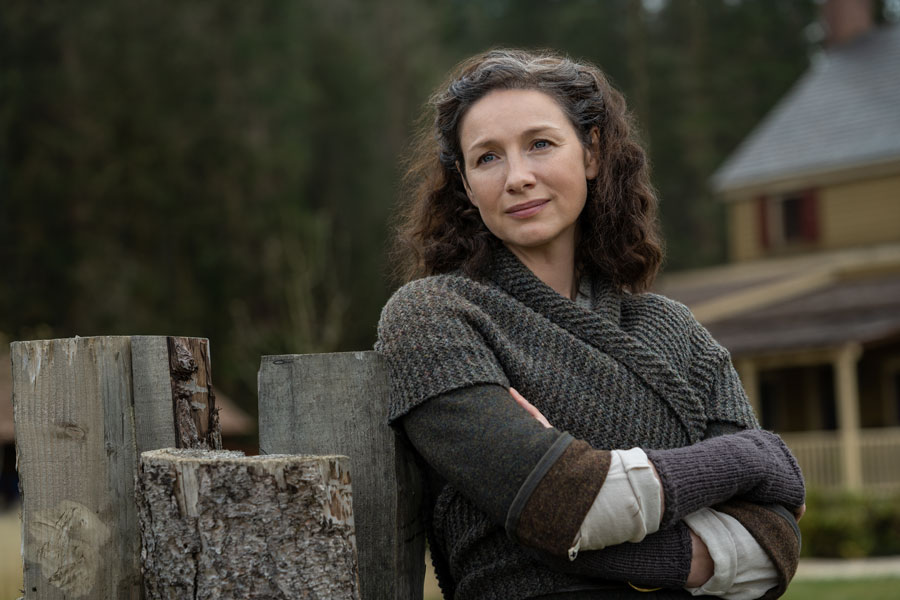 Woman with curly hair wearing layered knit sweaters and gloves, leaning against stacked firewood outdoors near a yellow house.
