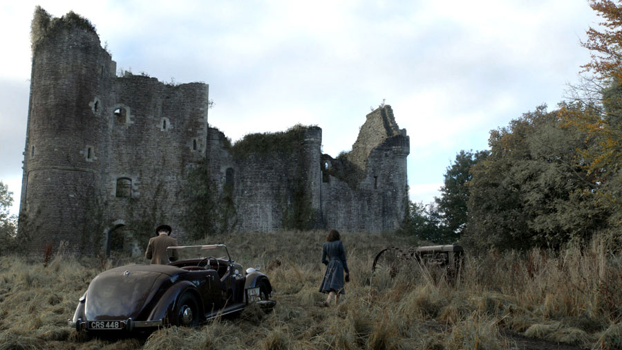 Two people near an old convertible car and rusty tractor in front of a large stone castle ruin surrounded by dry grass and trees.